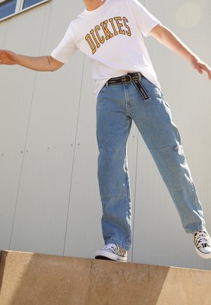 Person wearing white Dickies t-shirt, blue jeans, and zebra-patterned sneakers balancing on a concrete ledge outdoors.