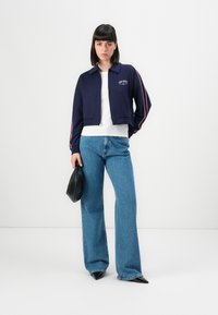 Crop navy jacket with white and burgundy stripes, zip front, worn over a white top, paired with high-waisted blue jeans and a black bag.