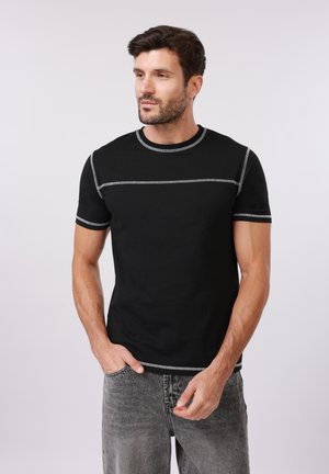 Άνδρας με γενειάδα, φοράει μαύρο πουκάμισο με κοντά μανίκια και λευκό ραφτό και γκρι τζιν, στέκεται με το ένα χέρι στην τσέπη, κοιτώντας προς τα αριστερά.