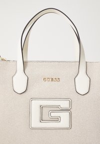 Beige canvas shoppingväska med två vita läderremmar. Har en framträdande vit G-logotyp och guld "GUESS"-branding. Slät textur.