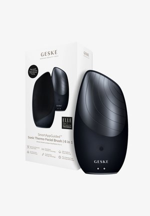 Zwarte Sonic Thermo Gezichtsborstel met geëxpandeerde siliconen haartjes, gladde ronde oppervlakte en bedieningsknoppen, naast witte verpakking met merk.