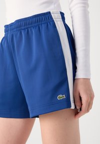 Calções de desporto azuis, feitos de tecido leve, com riscas brancas nas laterais e um pequeno logotipo de crocodilo verde na bainha inferior.