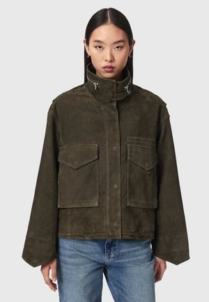 Femme portant une veste oversize en daim vert olive foncé avec de grandes poches à rabat et un col haut, associée à un jean bleu clair, fond neutre.