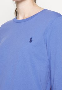 T-shirt violet à manches longues en coton doux, arborant un petit logo noir brodé sur la poitrine gauche et un col rond.