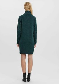 Robe en tricot verte avec un col montant et des manches longues, présentant une coupe ajustée se terminant au-dessus du genou, associée à des bottines noires à talons carrés.