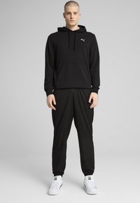 Sort hættejakke med kængurulomme og snøre, parret med løse sorte joggers. Hvide sneakers med grønne detaljer fuldender outfittet.