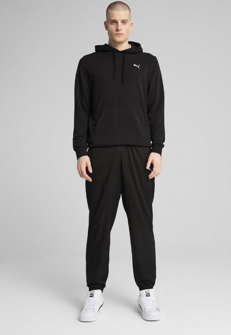 Sort hættejakke med kængurulomme og snøre, parret med løse sorte joggers. Hvide sneakers med grønne detaljer fuldender outfittet.
