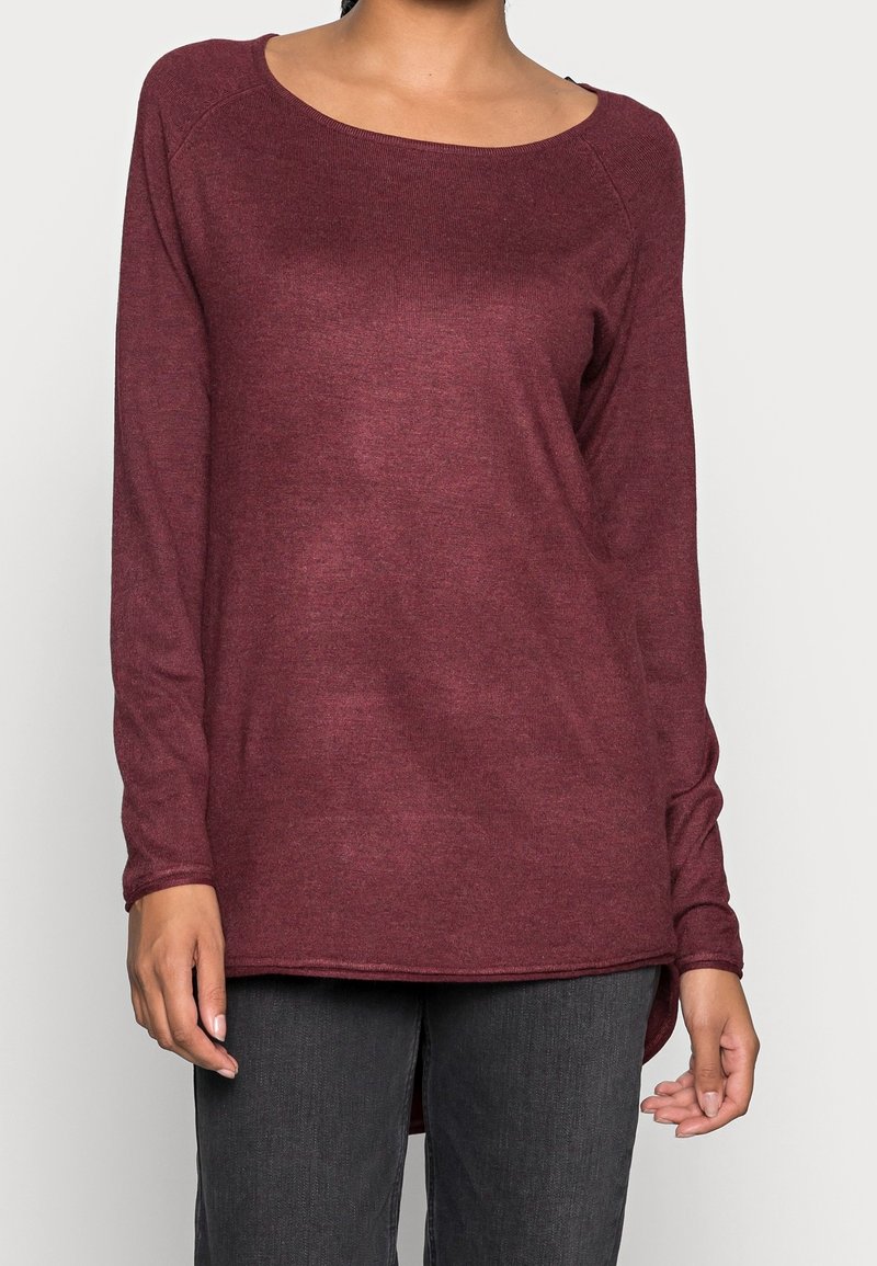 Kobieta ubrana w długą, bordową dzianinową bluzkę z szerokim, okrągłym dekoltem oraz ciemnoszare spodnie, stojąca na jednolitym tle.