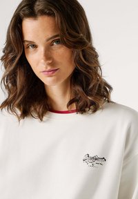 Biela mikina s okrúhlym výstrihom, s malým čiernym logom na hrudi. Mäkká textúra a ľahká látka s voľným strihom.