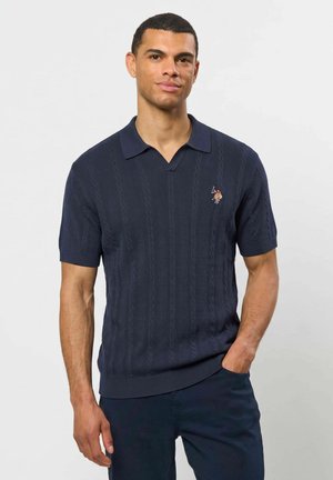 Homme portant un polo bleu marine à manches courtes avec un motif texturé et un petit logo brodé sur la poitrine, debout avec une main dans la poche.