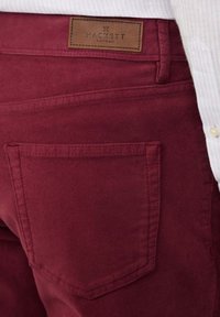 Bordeaux katoenen broek met een gladde textuur, voorzien van achterzakken en een leren label met de opdruk "HACKETT LONDON."