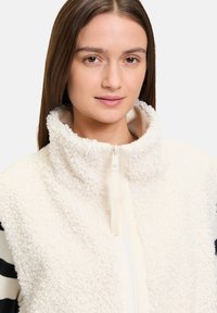 Gilet di pile bianco con colletto alto, chiusura con zip e superficie testurizzata. Presenta maniche a strisce bianche e nere. Morbido, caldo e leggero.