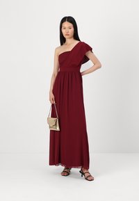 Rochie maxi burgundă fără bretele cu un corsaj din dantelă și detalii asimetrice la umăr, asortată cu o poșetă aurie și sandale negre.