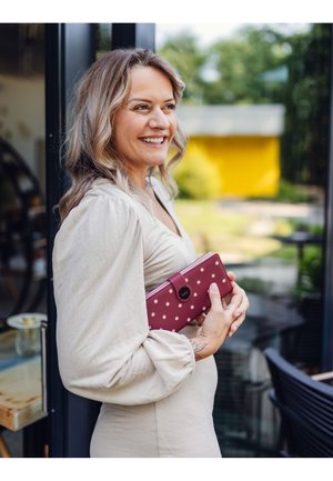 Magenta wallet with white polka dots, rectangular shape, secured with a black button closure. Woman in beige dress holds wallet against her body.