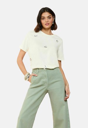 Cremfarbenes Kurzarm-T-Shirt mit Schmuckverzierungen; kombiniert mit hochgeschnittenen, hellgrünen, weiten Hosen. Glatte Stofftextur.