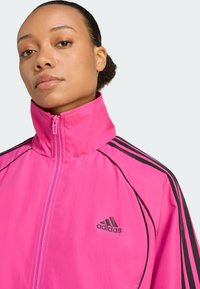 Adidas pink zip-up jakke med høj krave, med sorte striber langs ærmerne og et lille sort logo på brystet. Glat tekstur.