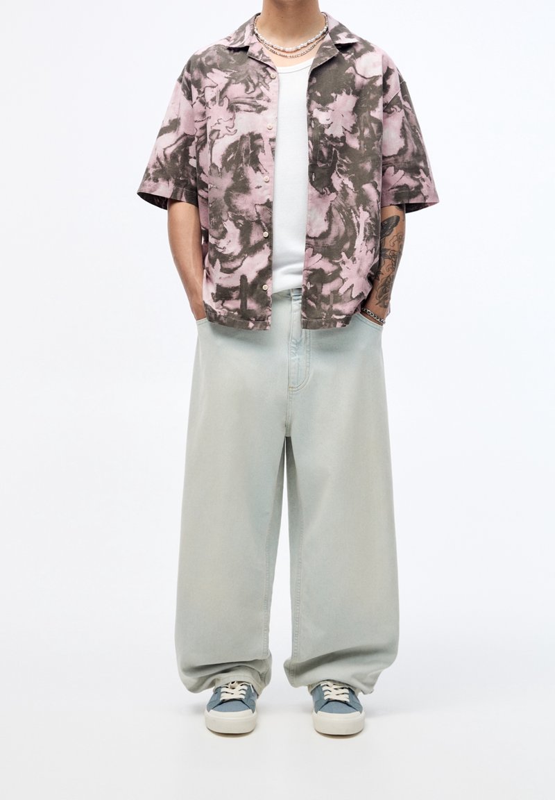 Camicia a maniche corte, a righe rosa e grigia, abbinata a jeans palazzo verde chiaro e sneakers con dettagli blu. Sotto indossa una semplice t-shirt bianca.