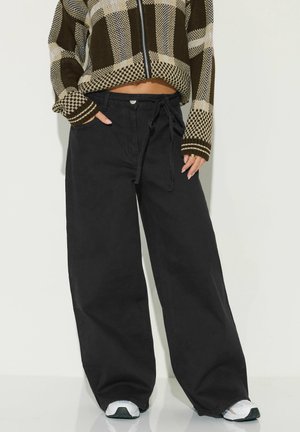 Person iført sorte bukser med vide ben, snøre og knap, cropped brun ternet lynlås-sweater og hvide sneakers, hænder i lommerne.