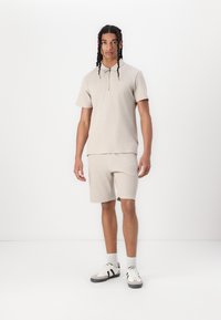 Beige strik polo med kortærmer og halvlængde lynlås, parret med matchende shorts. Modellen har grå og hvide træningssko på med sokker.