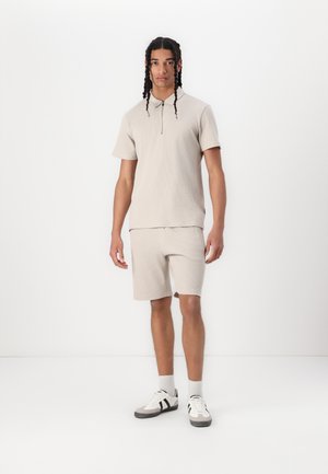 Beige pletené polokošeľa s krátkym rukávom a polovičným zipsom, skombinovaná s krátkymi nohavicami v rovnakej farbe. Model má na sebe šedé a biele športové topánky so ponožkami.