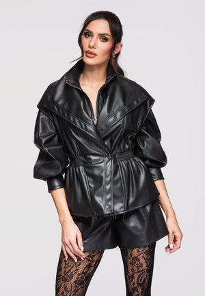 Donna che indossa una giacca di pelle nera con maniche a sbuffo, pantaloncini di pelle abbinati e calze nere con motivo all'uncinetto, in piedi davanti a uno sfondo semplice.