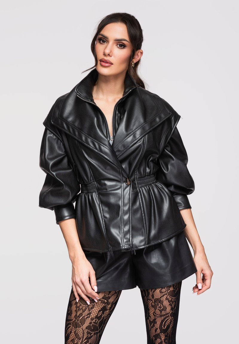 Mujer vistiendo chaqueta de cuero negra con mangas abullonadas, pantalones cortos de cuero a juego y medias negras con estampado de encaje, posando frente a un fondo liso.