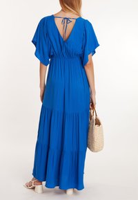Robe maxi bleue avec des manches volantées, taille froncée et jupe à volants. Design dos nu avec un détail à nouer. Le mannequin tient un sac en osier.
