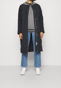 Manteau d'extérieur noir matelassé avec des fermetures à pression, porté sur un hoodie gris, associé à un jean bleu à jambes larges et des baskets blanches.