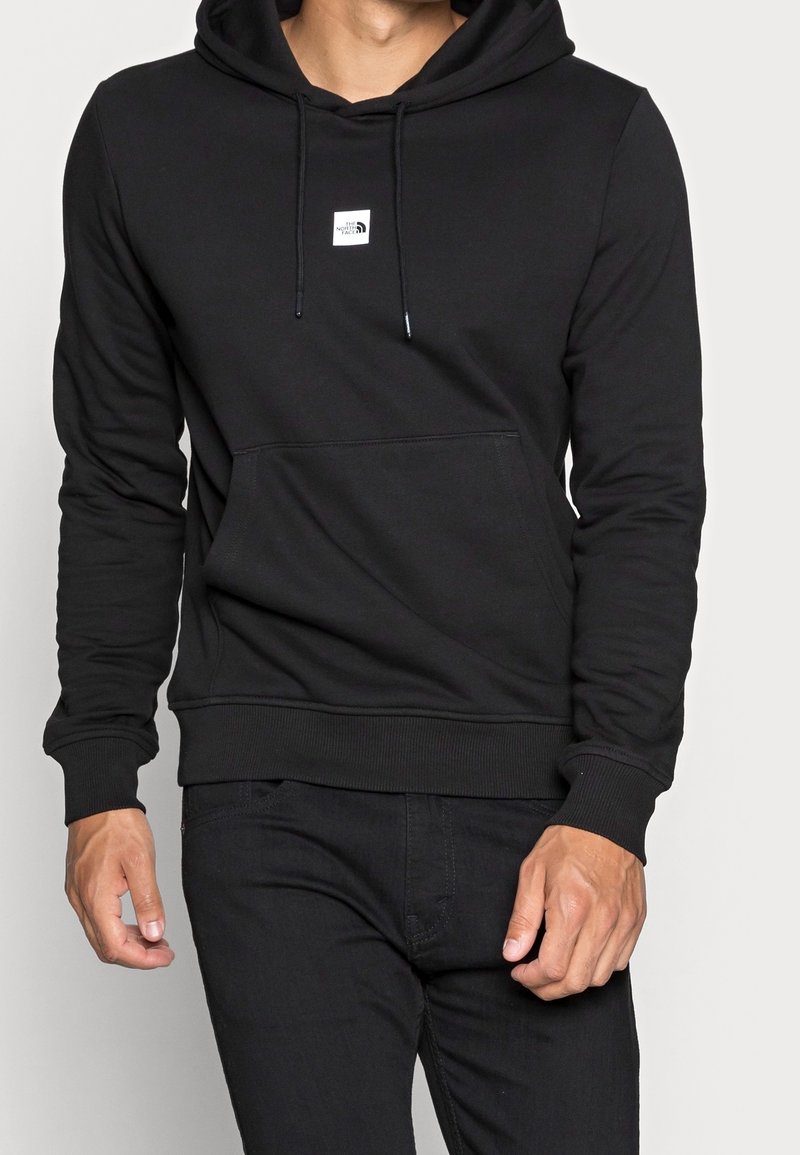 Homme portant un sweat à capuche noir avec une poche avant et un petit logo blanc North Face, associé à un pantalon noir.