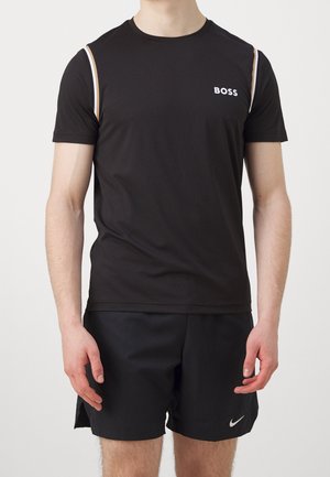 Mężczyzna noszący czarną koszulkę BOSS z białym obszyciem na ramionach i czarne krótkie spodenki Nike, stojący na jednolitym tle.