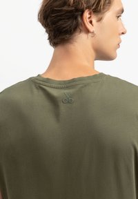 Oliegroene t-shirt met ronde hals, gemaakt van zachte stof. Heeft een kleine schaarafbeelding geborduurd op de achterkant.