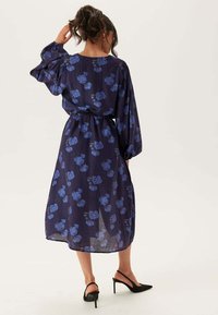 Kvinna med mörkt hår i en knut som bär en marinblå blommig midiklänning med långa ärmar och svarta högklackade slingback-skor.