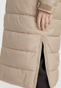 Beige puffjacka med sydd panelering, en sidzipficka och stängning med kardborreband vid ärmarna, med en slät, matt yta.