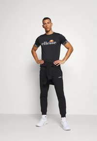 Svart kortärmad t-shirt med logga, kombinerad med svarta joggers. Båda är gjorda av mjukt tyg. Modellen bär vita sportskor.