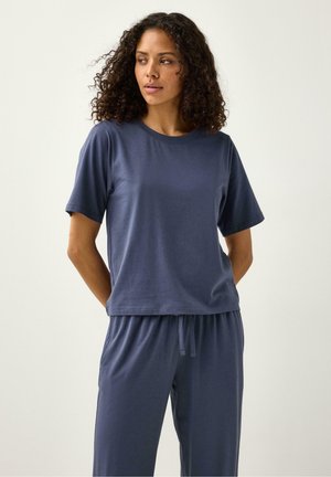 Kvinde med krøllet hår iført løs, marineblå t-shirt med korte ærmer og matchende bukser med snøre, stående med hænderne bag ryggen.