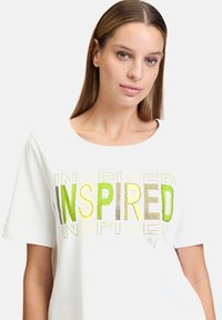T-shirt en coton blanc avec un graphique audacieux "INSPIRED" en couleurs vertes et métalliques, avec un col rond et des manches courtes.