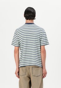 Stribet T-shirt i lys grøn, navy og hvid; korte ærmer, rund halsudskæring, blødt stof; båret med lyse beige bukser, set fra ryggen.