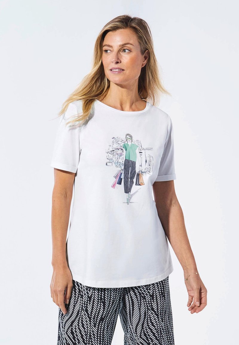 Weißes T-Shirt mit kurzen Ärmeln, das ein farbenfrohes Grafikmotiv einer Person beim Einkaufen zeigt, kombiniert mit weiten schwarz-weißen gemusterten Hosen.