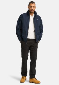 Marineblauwe lichtgewicht jas met een opstaand kraag, witte polo eronder, zwarte broek en tan kleuren laarzen. Bevat een logo op de borst.