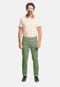 Camisa polo de manga corta con un patrón floral en tonos rosas, con un cuello rojo, combinada con pantalones verdes. El modelo, que lleva zapatos negros, se presenta en posición erguida.