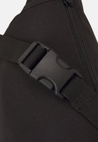 Czarna torba materiałowa z bocznym zamkiem błyskawicznym i regulowanym paskiem z teksturowaną plasticzną klamrą. Zbliżenie podkreśla materiały i design.