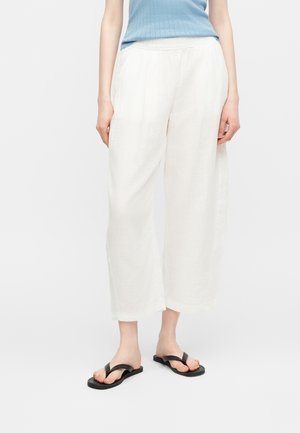 Osoba oblečená v voľných bielych nohaviciach, čiernych žabkách a svetlomodrom tielku bez rukávov, stojaca na bielom pozadí.