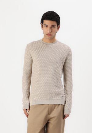 Maglione a maglia beige con texture, scollo rotondo e polsini a costine. Presenta un motivo a tessuto simile a una griglia e un logo laterale. Abbinato a pantaloni chiari.