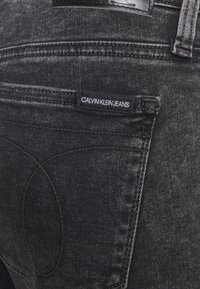 Jeans de mezclilla negra con una textura suave, que presentan costuras en contraste, una etiqueta en el bolsillo trasero con la inscripción "CALVIN KLEIN JEANS" y un diseño ajustado.