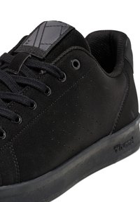 Baskets en daim noir avec un design perforé, des lacets ronds et une semelle en caoutchouc noir texturé. La languette présente un logo subtil et un motif.