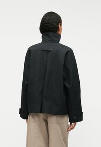 Chaqueta negra, ligera, con cuello alto, puños de botón y corte cuadrado. Presenta una etiqueta de logo en la parte posterior, textura suave y diseño minimalista.