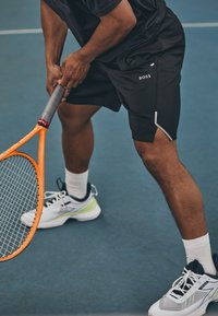 Pantalones cortos deportivos negros con un detalle de raya blanca, combinados con zapatillas de tenis blancas y azul marino. Se sostiene una raqueta de tenis con un marco naranja.