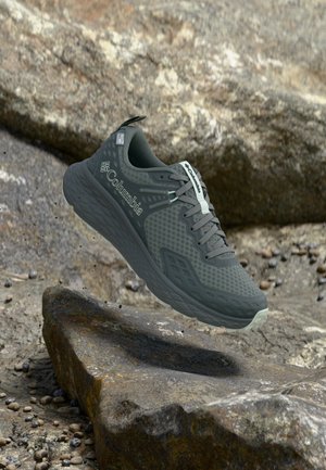 Chaussure de randonnée Columbia gris foncé flottant au-dessus d'un terrain rocheux avec une semelle texturée et une tige en mesh pour la respirabilité.