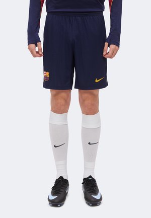 Homme portant un short de football FC Barcelone bleu foncé, des chaussettes blanches montantes Nike et des crampons noirs Nike, debout sur un fond blanc.