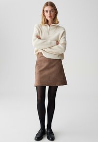 Cremfarbener gestrickter Pullover mit einem Reißverschlusskragen, kombiniert mit einem braunen Wildleder-Minirock und schwarzen, durchsichtigen Strumpfhosen, abgerundet mit schwarzen Slippern.